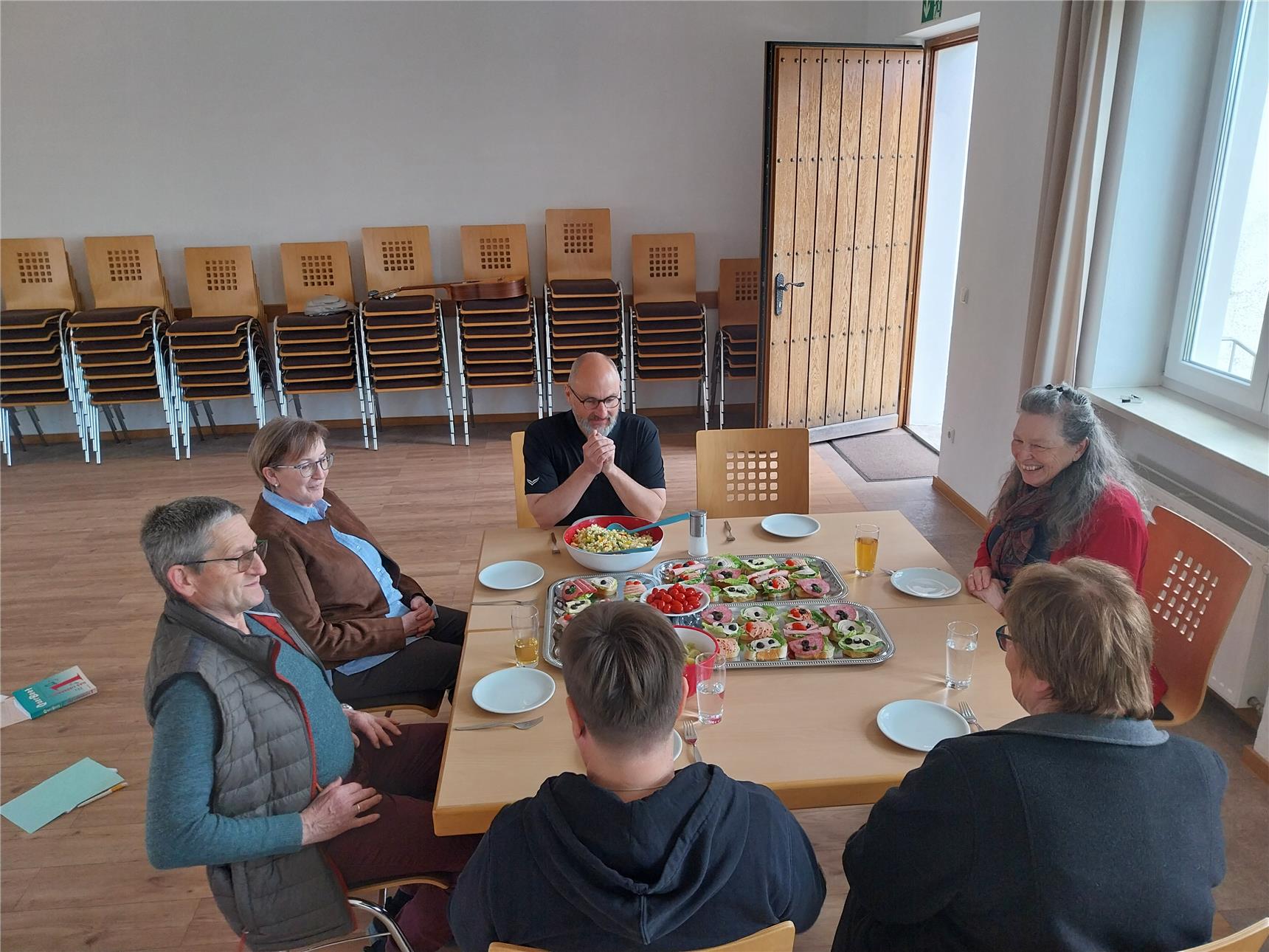 Sechs Personen sitzen fröhlich um einen Tisch mit belegten Broten, Rohkost und Getränken. Im Hintergrund sind gestapelte Stühle und eine geöffnete Holztür zu sehen.
