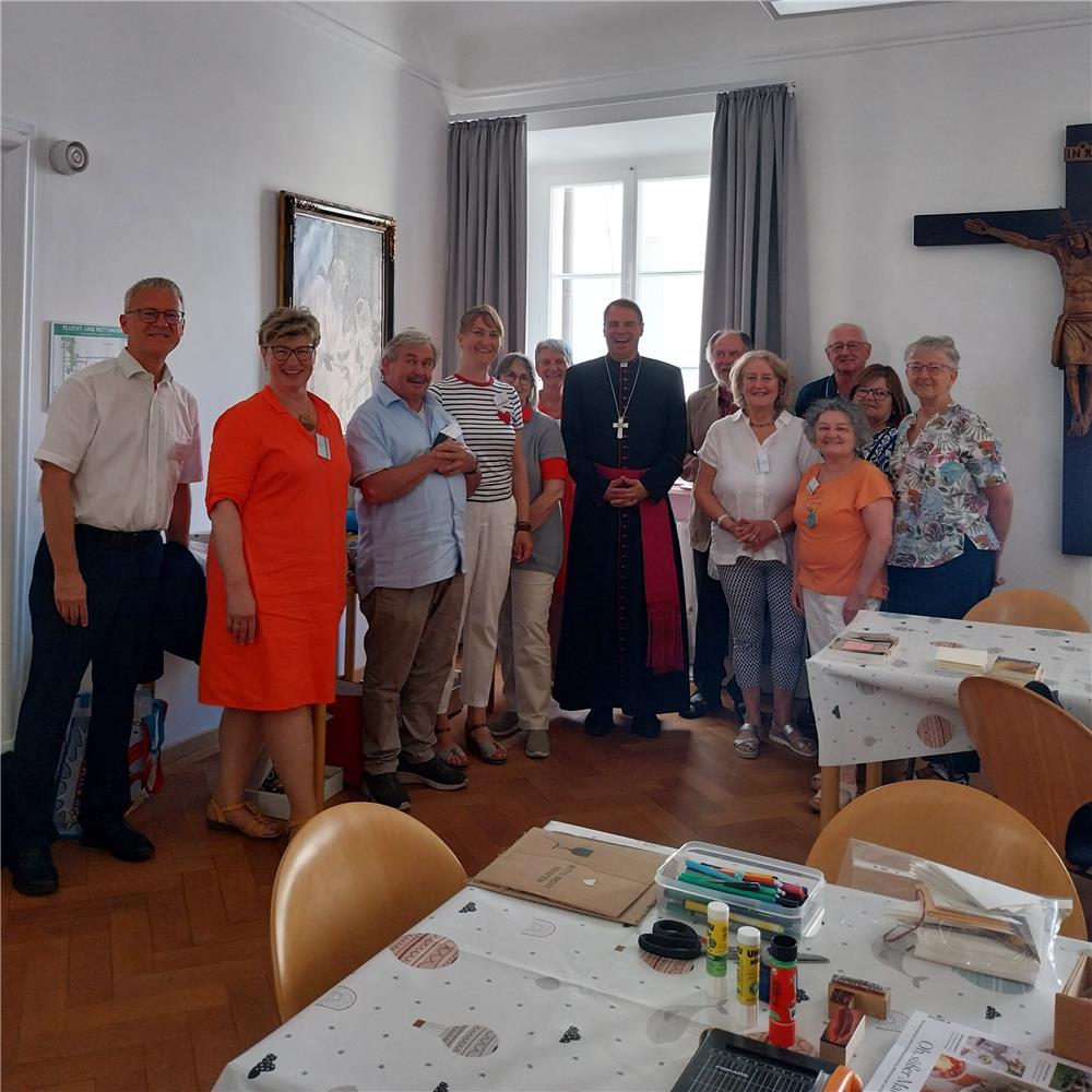 Gruppe von Menschen in einem hellen Raum, die gemeinsam mit Bischof Stefan Oster für ein Gruppenfoto posieren. Im Vordergrund liegen Bastelmaterialien wie Kleber, Stifte und Stempel auf Tischen mit gepunkteten Tischdecken.