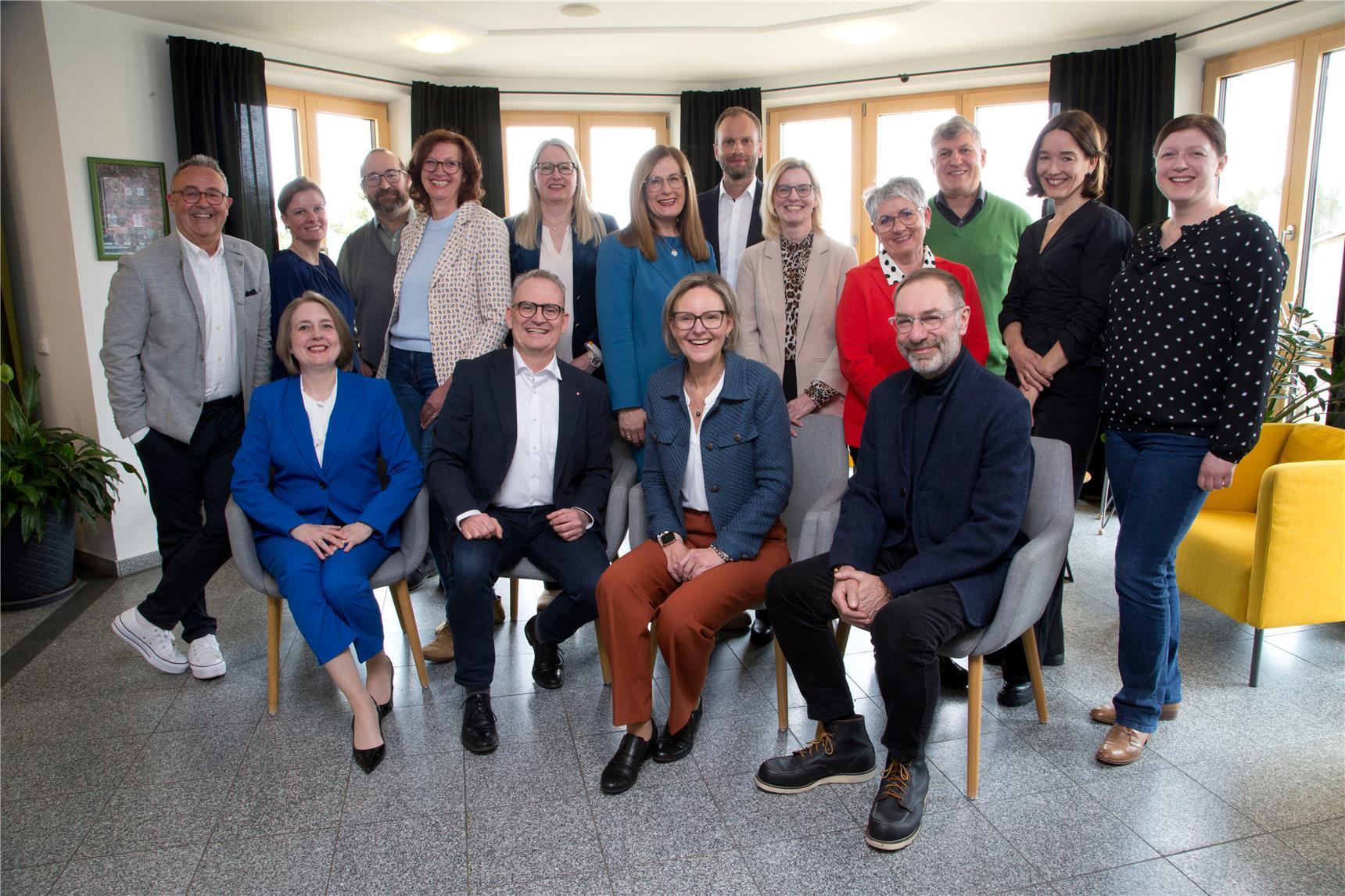 Gruppenfoto von fünfzehn Personen, die in einem hellen Raum mit großen Fenstern und gelben Sesseln posieren – drei sitzen vorn, während die restlichen im Halbkreis dahinter stehen und lächeln.