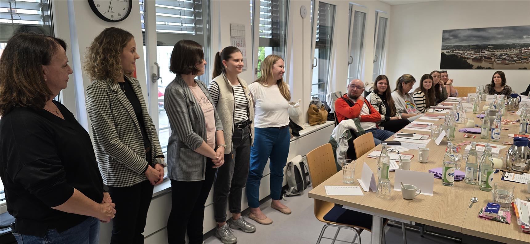Eine Gruppe von Frauen steht nebeneinander vor einem Fenster, während weitere Personen an einem langen Konferenztisch sitzen und zuhören.