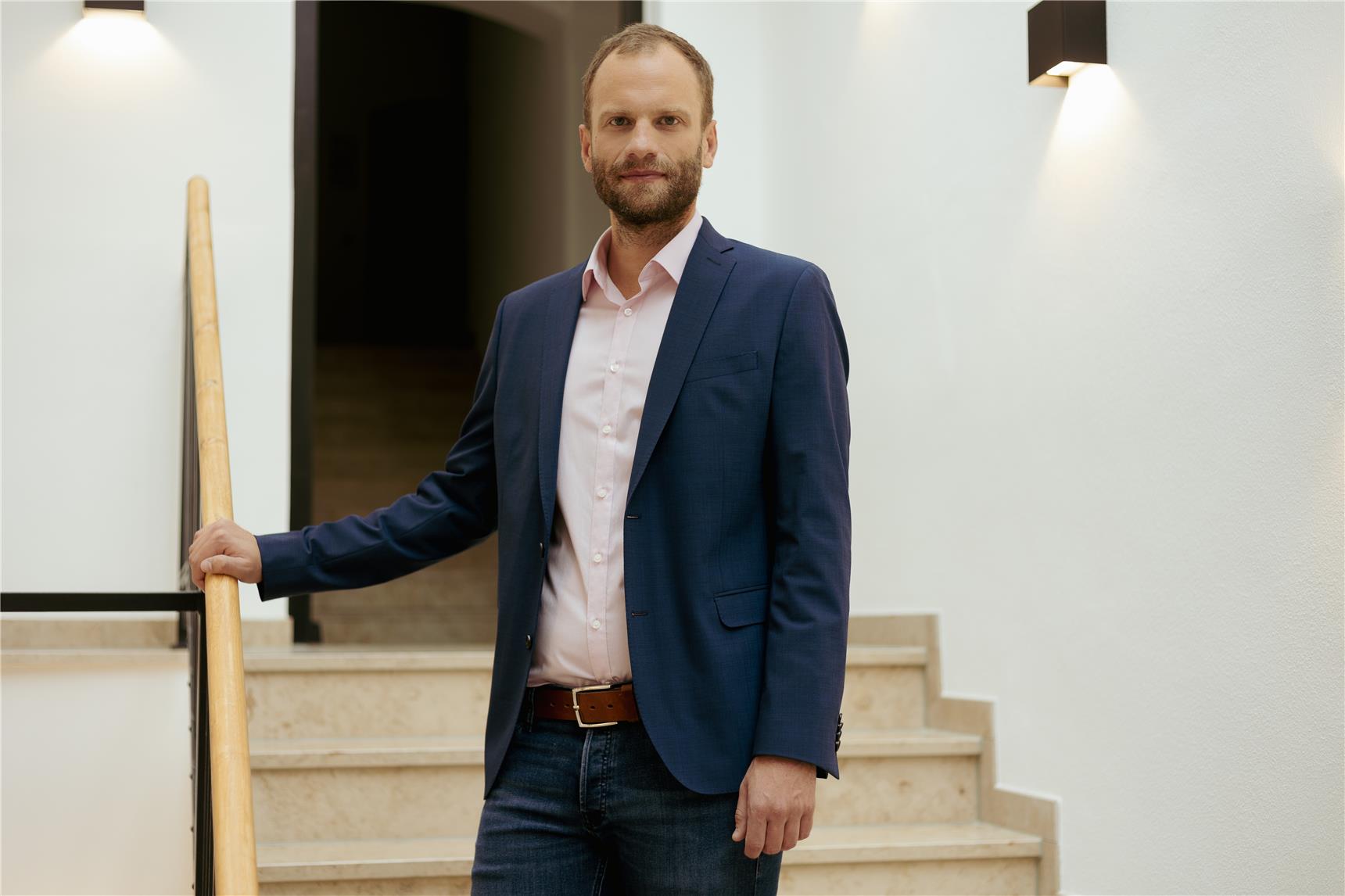 Personalleiter Thomas Furthmeier - Ein Mann mit kurzem Bart und kurzem Haar steht auf einer Treppe in einem modernen, hellen Gebäude. Er trägt ein dunkelblaues Sakko, ein hellrosafarbenes Hemd und eine Jeans und hält sich locker am Geländer fest.