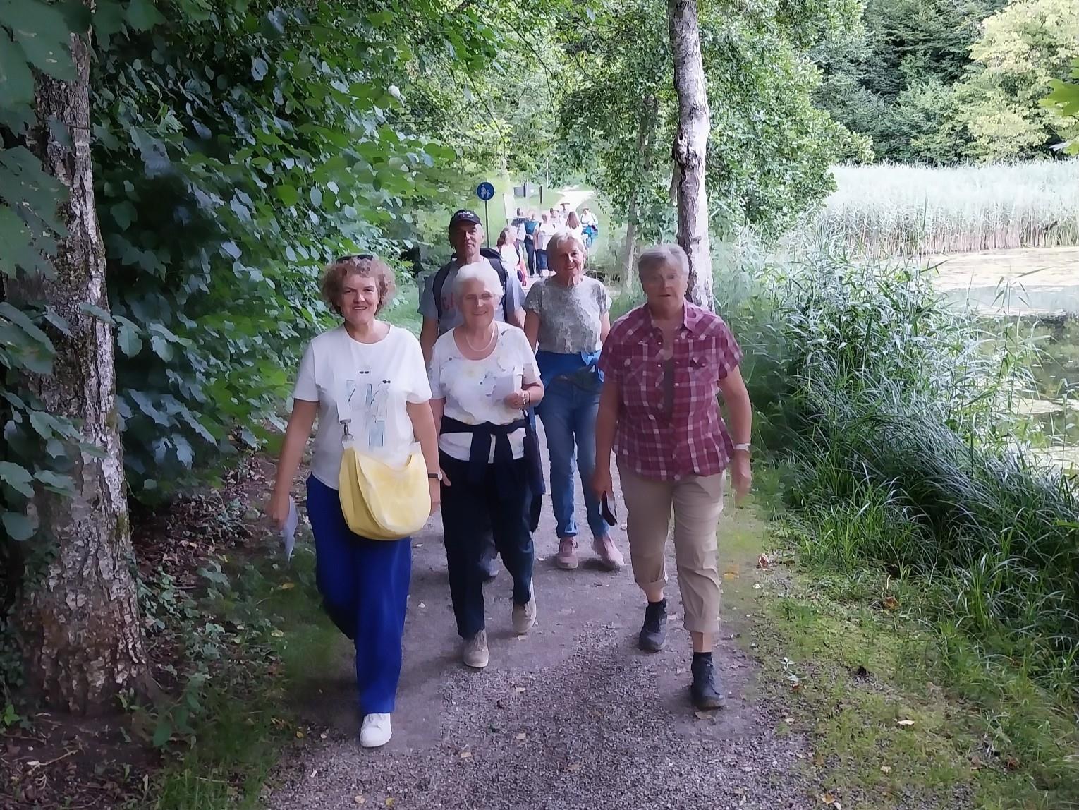 Eine Gruppe wandert lächelnd auf einem schattigen Waldweg entlang eines Teiches. Die Stimmung wirkt entspannt und gemeinschaftlich.