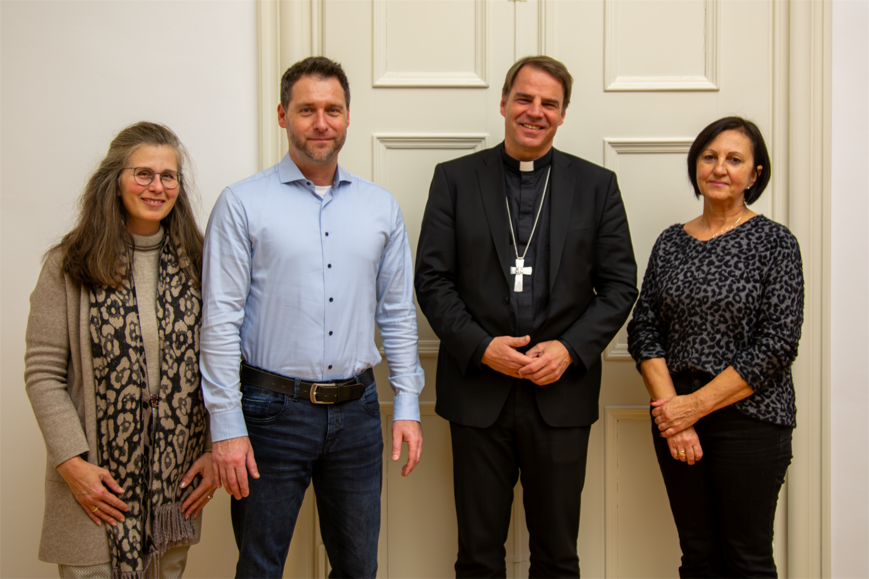 Das Bild zeigt vier Personen, die nebeneinander vor einer hellen Tür stehen. Die zweite Person von rechts ist ein Geistlicher in schwarzer Kleidung mit einem Kreuz um den Hals. Die anderen drei Person