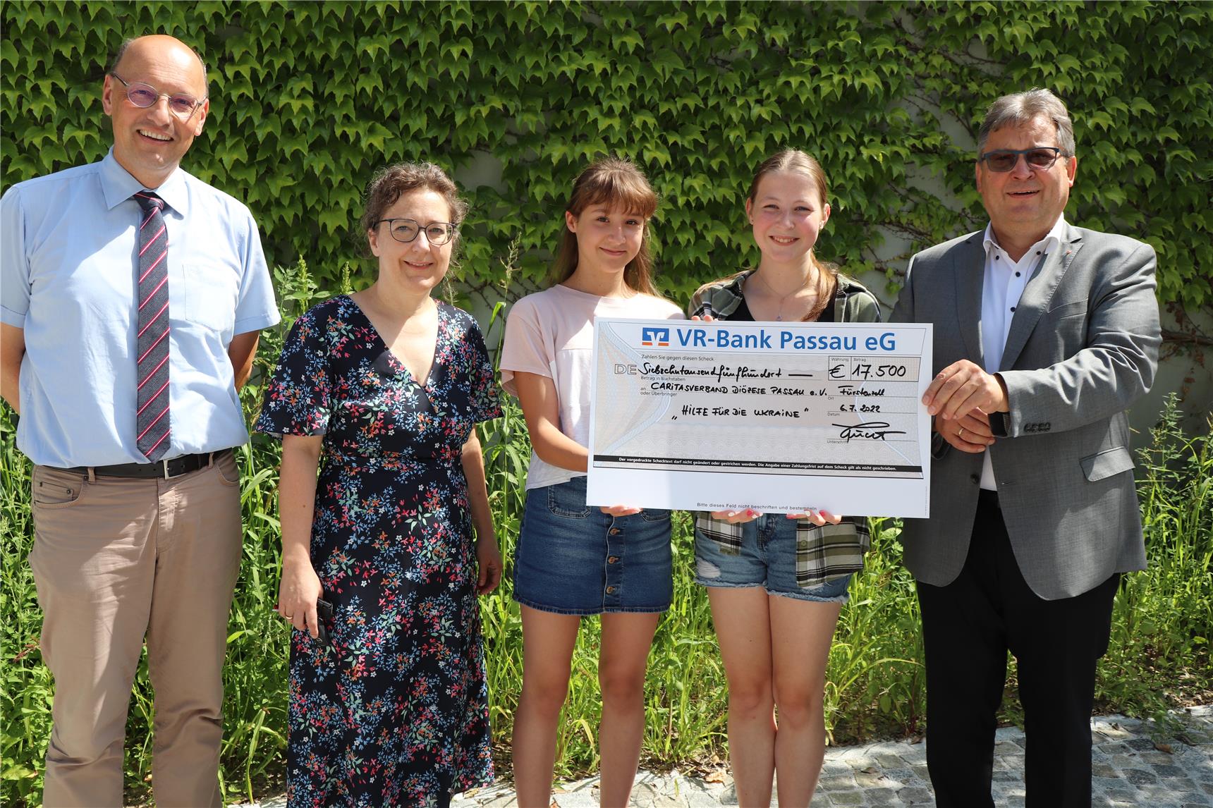 2 Schülerinnen, 1 Lehrerin, Caritas-Diakon Konrad Niederländer und Schulleiter OStD i.K.  Dr. Roland Feucht vor einer efeubewachsenen Wand präsentieren den Scheck