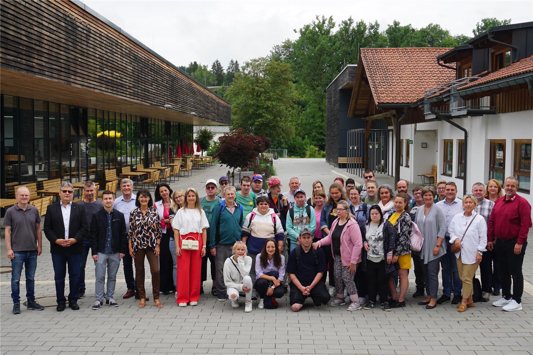 Eine große Gruppe von Menschen, darunter Männer und Frauen unterschiedlichen Alters, posiert für ein Gruppenfoto vor einem modernen Gebäude mit großen Fenstern und Holzverkleidung. 