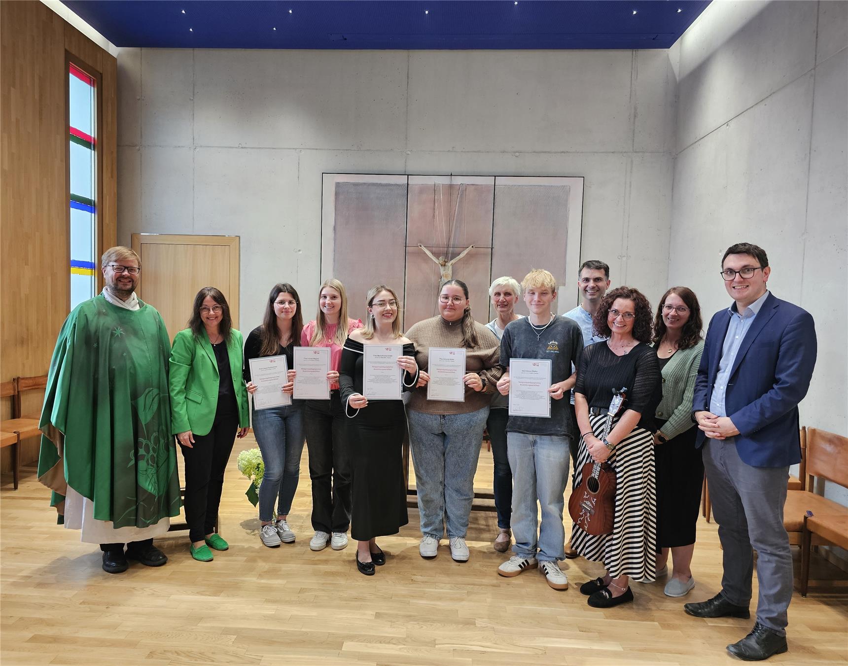 Gruppenfoto von zwölf Personen in einer Kirche, fünf davon halten Zertifikate in der Hand. Im Hintergrund hängt ein modernes Kruzifix an der Wand.