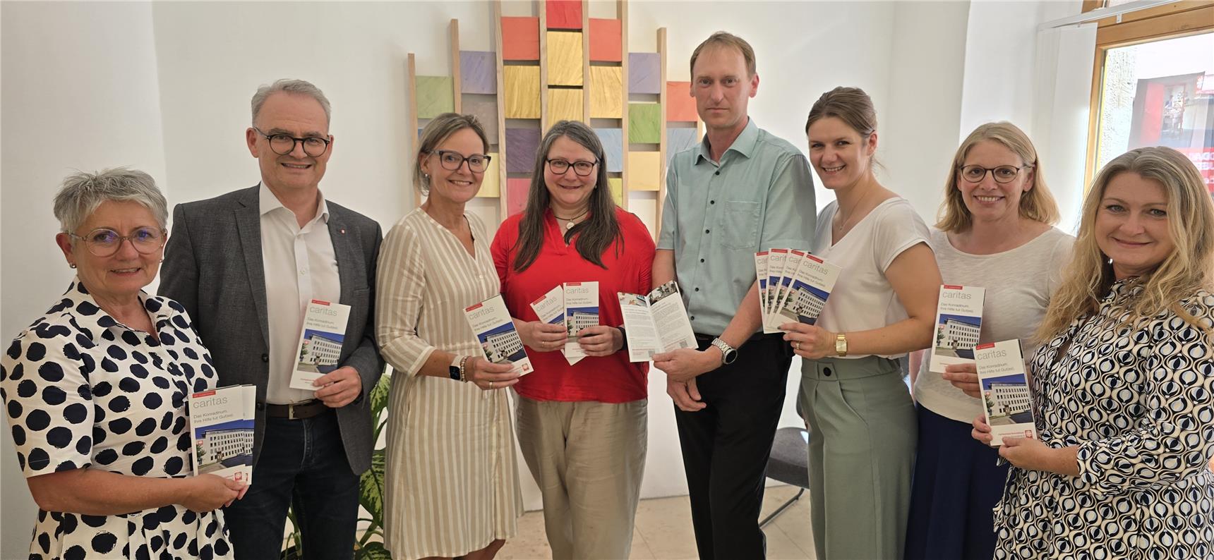 Das Bild zeigt acht lächelnde Erwachsene, die nebeneinander stehen und Broschüren der Organisation “Caritas” in den Händen halten. Die Broschüre trägt den Titel „Das Caritas Zentrum stellt sich vor“, mit einem Foto des Gebäudes auf dem Cover. Im Hintergrund hängt eine farbenfrohe Wanddekoration aus bunten, rechteckigen Holzplatten.
