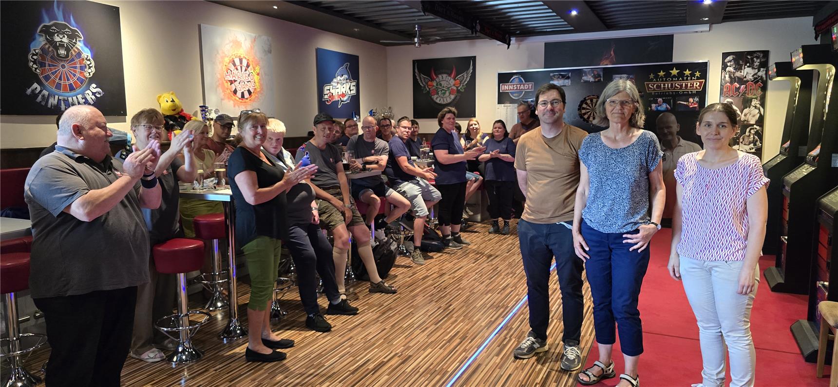 Viele Personen sind in einem Raum, der mit mehrere Dartscheiben ausgestattet ist. Alle lachen in die Kamera. Der Raum ist mit auffallenden Bildern an den Wänden dekoriert.