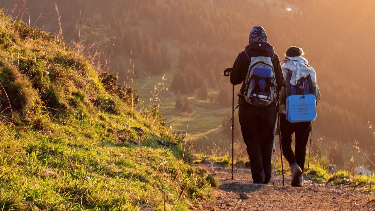 Zwei Pilger mit Rucksäcken und Wanderstöcken wandern bei Sonnenuntergang auf einem schmalen Pfad durch die Berge. Das warme Licht taucht die weite, bewaldete Landschaft in eine friedliche Stimmung.