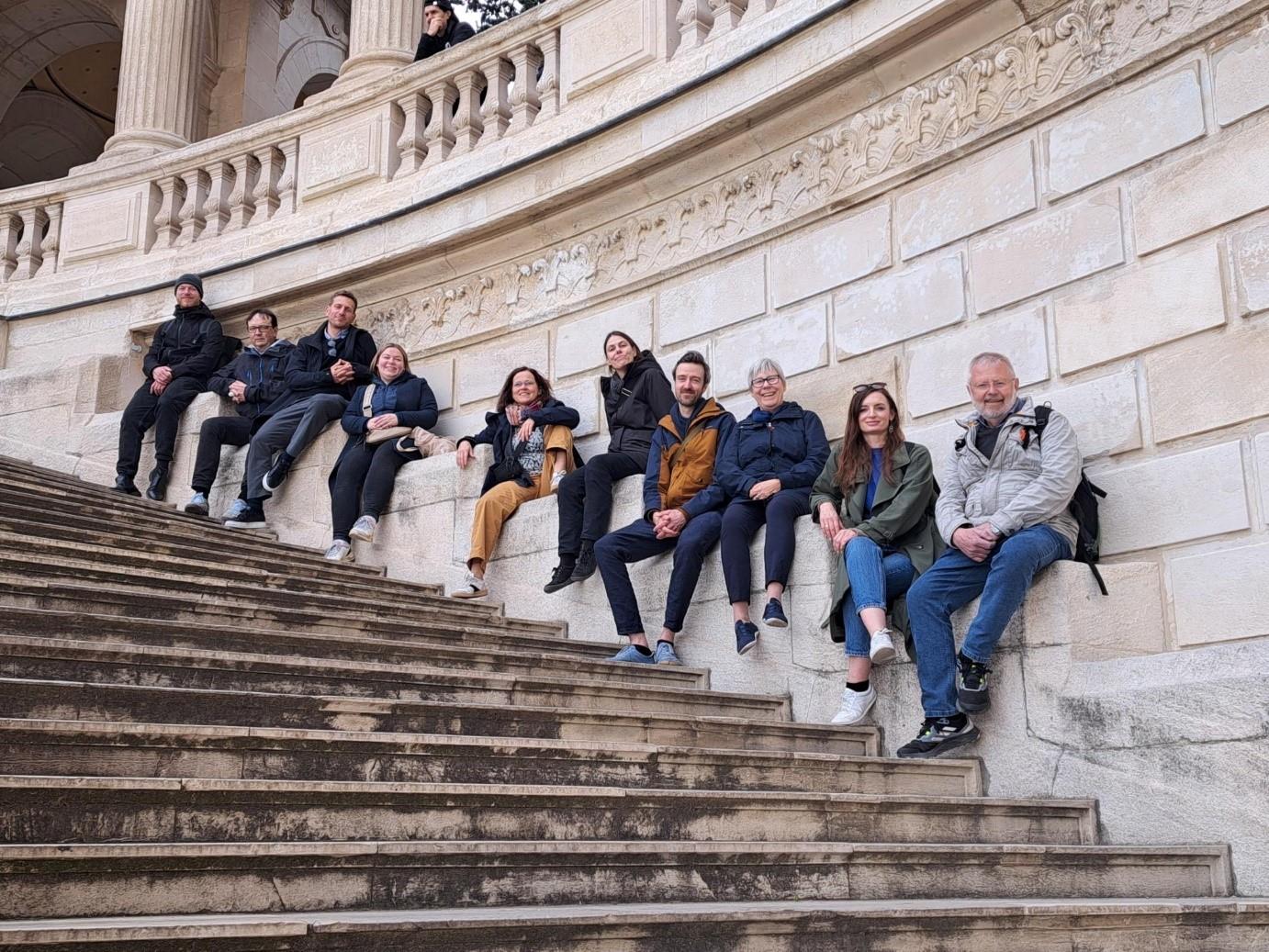 10 Personen sitzen verteilt auf steinigen Treppenstufen entlang einer alten steinigen Treppe. Alle lachen in die Kamera.