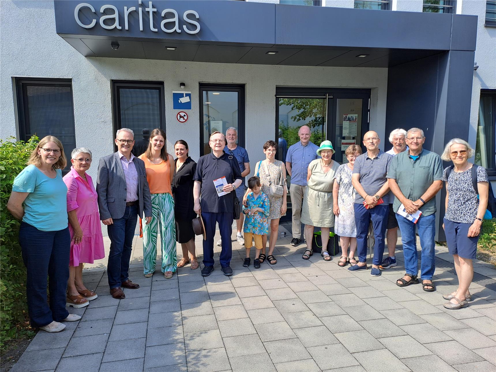 Etwa 16 Personen stehen vor einem weißen Gebäude der Caritas. Alle lachen in die Kamera.