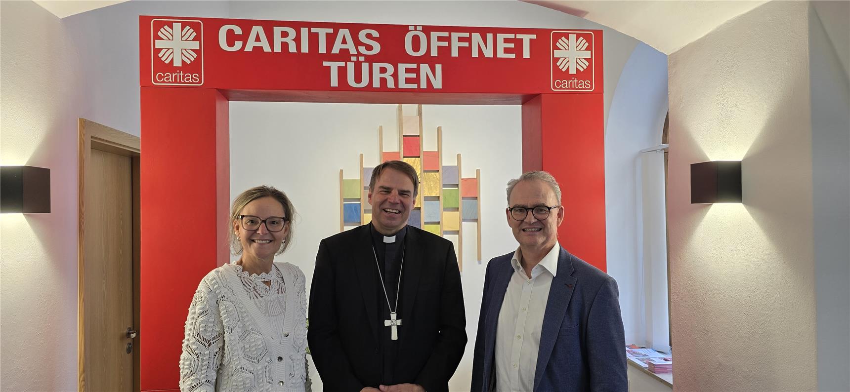 Drei Personen stehen vor einem roten Caritas-Bogen. Auf dem Bogen steht: 