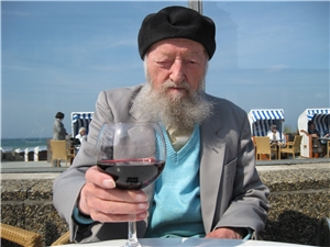 Ein alter Mann mit langem grauen Vollbart und Mütze hat ein Glas Wein in der Hand, im Hintergrund die Ostsee.