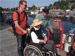 Ein ehrenamtlicher Helfer mit einem alten Herrn und einer Bewohnerin im Rollstuhl am Hafen von Eckernförde.