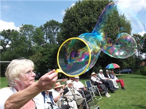 Eine Betreuerin produziert Riesenseifenblasen im Park, Bewohner sitzen auf Campingstühlen in der Sonne und schauen zu.