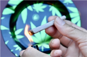Closeup: Anzünden eines Joints. Im Hintergrund unscharf ein Aschenbecher mit Cannabisblättermuster in grün und schwarz.