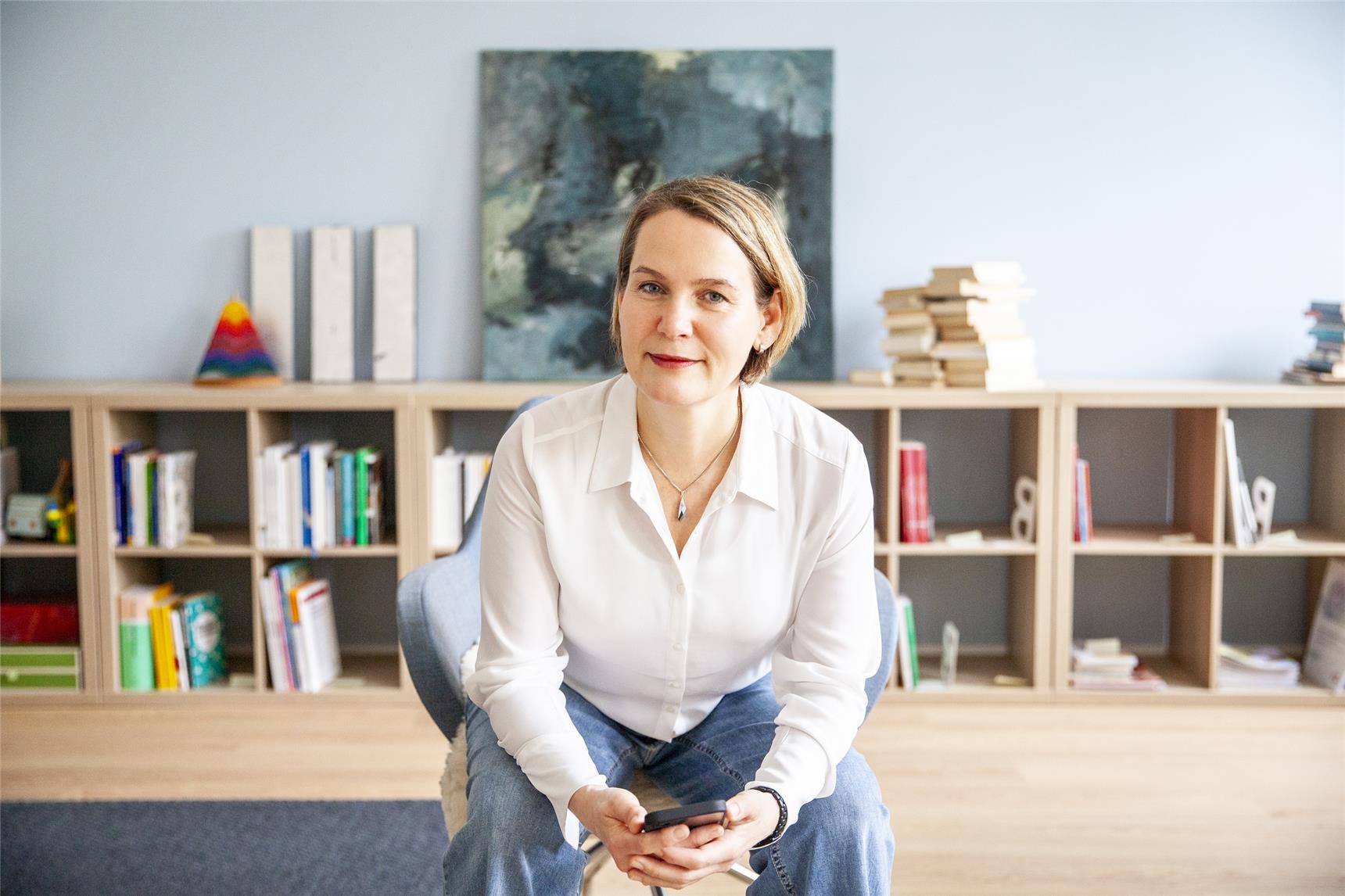 Delija sitzt in ihrem neuen Büro in Regensburg vor einem kniehohen Bücherregal und pastellblauer Wand