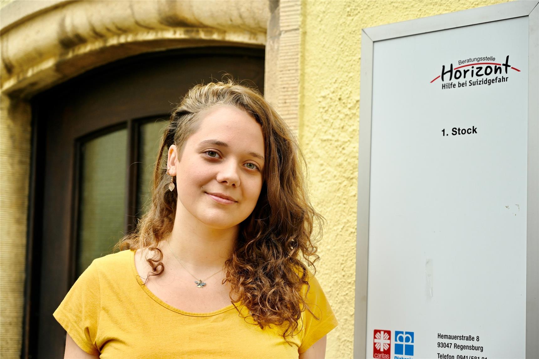 Porträt von Lisa Pfeiffer. Sie trägt lange, hellbraune Haare und ein gelbes Shirt. Sie steht vor ihrer Beratungsstelle Horizont.