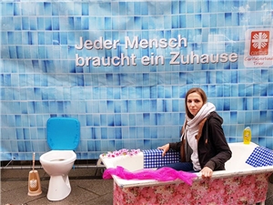Eine Frau sitzt in einer Badewanne; hinter ihr ein Transparent mit Badezimmerkacheln und der Aufschrift 