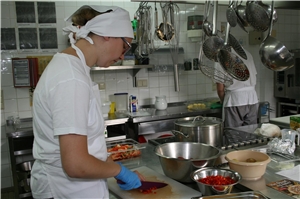 Zwei Personen in Berufskleidung in einer Küche; im Vordergrund schneidet eine von ihnen Paprika.