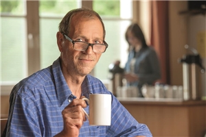 Ein Mann mit Brille hält eine Tasse und schaut in die Kamera; im Bildhintergrund: eine Frau an einer Getränketheke.