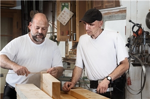 Zwei Männer mit Zollstöcken und Holzbalken in einer Werkstatt.