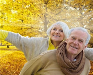 Im Vordergrund: ein alter Mann, dahinter eine alte Frau, die die Arme ausbreitet, beide lächeln; im Hintergrund: Herbstbäume in Gelb-Tönen
