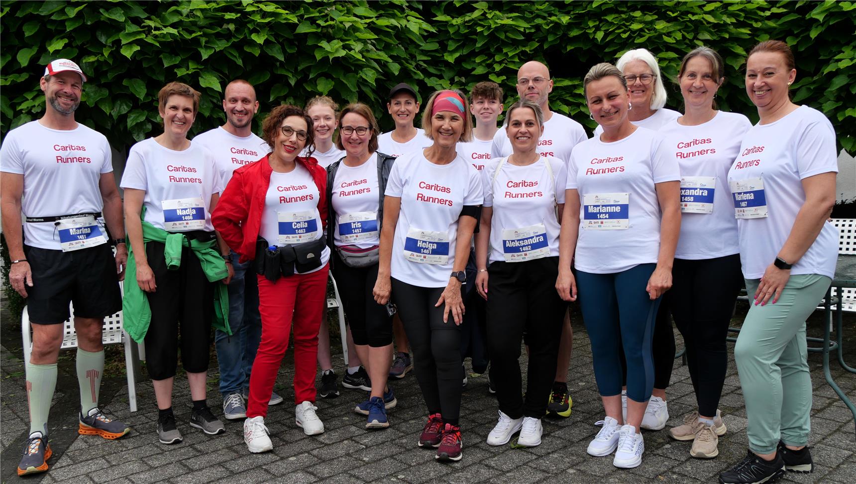 15 Frauen und Männer stehen in zwei Reihen hintereinander. Sie tragen Sportkleidung. Auf ihren weißen T-Shirts ist die Aufschrift 