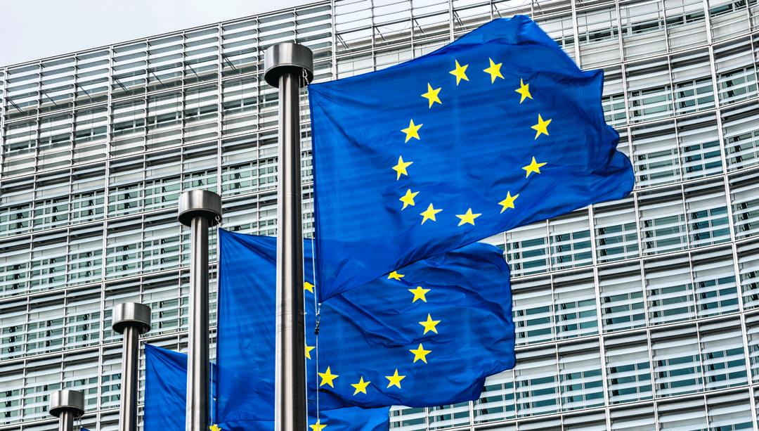 Drei EU-Flaggen wehen im Wind vor modernem Gebäude mit Glasfassade..