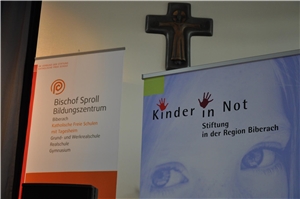 10 Jahre Stiftung Kinder in Not Banner der Stiftung Kinder in Not und des Bischof Sproll Bildungszentrum. Dahinter ein Kruzifix an der Wand