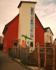 Ein rotes Haus mit einem weißen Vorbau, davor befindet sich ein umzäunter Kinderspielplatz.