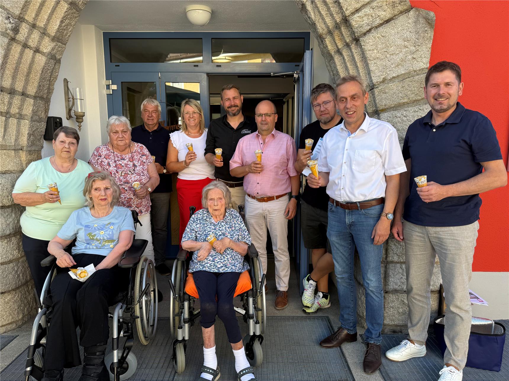 Gruppe von Senioren und Betreuern vor einem Gebäude, lächelnd und mit Eiswaffeln in der Hand; zwei ältere Frauen sitzen in Rollstühlen vorne, während der Rest der Gruppe stehend hinter ihnen posiert. Die Szene vermittelt eine freundliche Sommeratmosphäre mit einem Hauch von Gemeinschaftsgefühl.