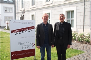 Professor Dr. Uwe Becker (links) und Weihbischof Franz Josef Gebert stehen vor dem Ausstellungsplakat "LebensWertArbeit".