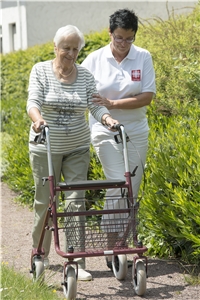 Eine ältere Frau mit ihrem Rollator wird von einer Pflegerin der Sozialstationen begleitet 