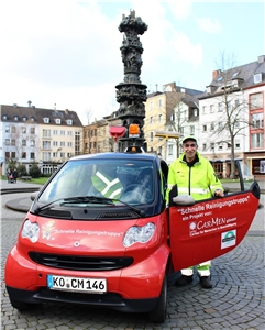 In der Koblenzer Innenstadt: Herr Duran steht fröhlich schauend neben seinem Auto, das die Aufschrift 'Schnelle Reinigungstrupps' trägt.