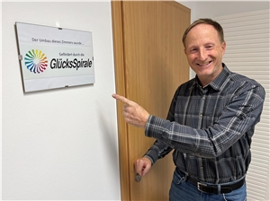 Joachim Grämer zeigt auf das Schild der GlücksSpirale, das zeigt, dass der Zimmerumbau gefördert wurde.