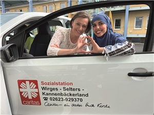 Bouchra und Halyna blicken durch das Fenster eines Autos der Caritas-Sozialstation und formen mit ihren Fingern gemeinsam ein Herz.