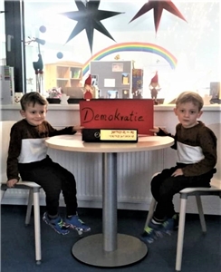  Julian und Nico vom Kinderparlament der Kita Regenbogen sitzen an einem Tisch und halten ein rotes Schild mit der Aufschrift Demokratie fest.