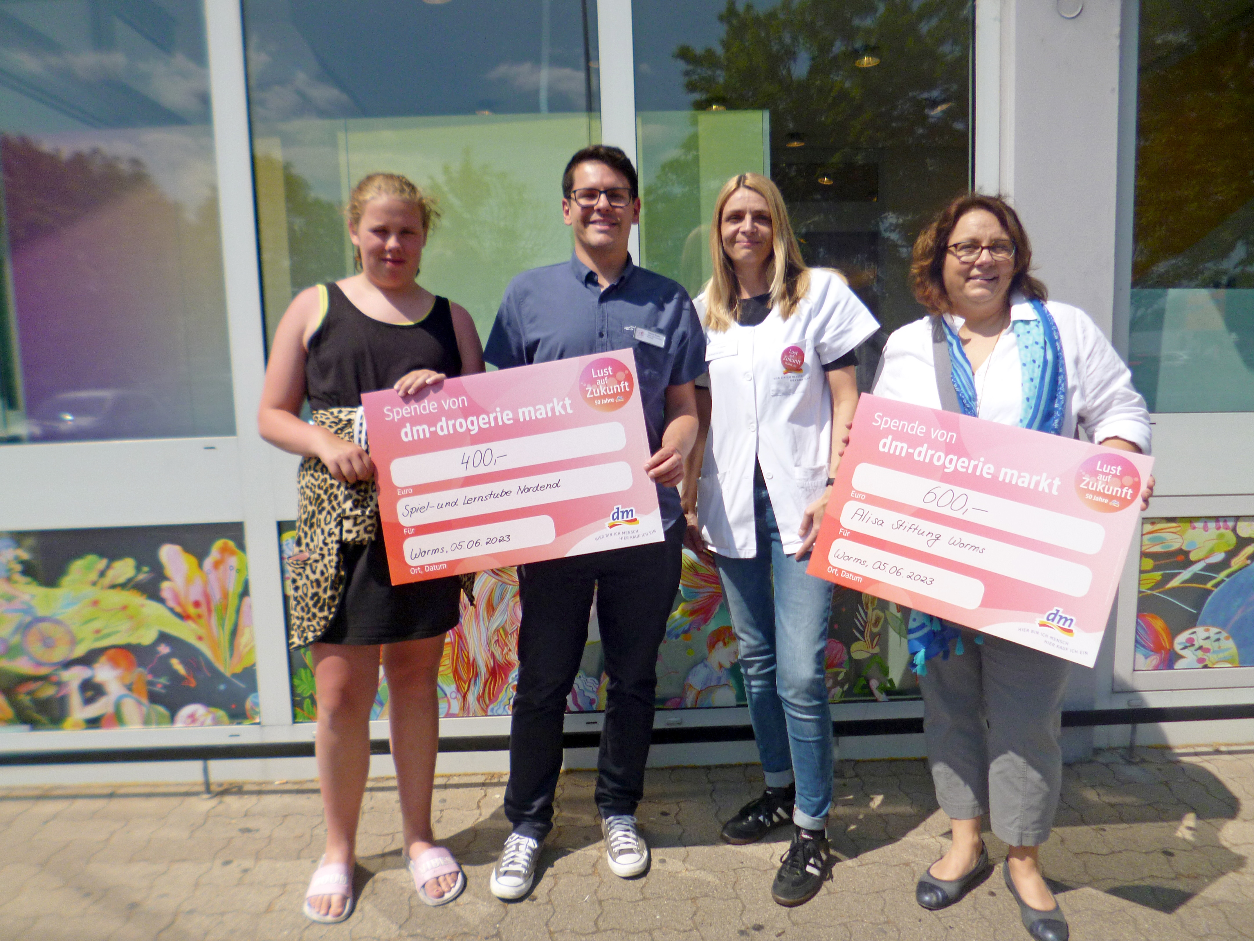 drei Frauen und ein Mann mit zwei Spenden-Schecks vor einem Geschäft