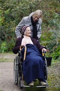 Junge Frau schiebt einen Rollstuhl in dem eine ältere Frau sitzt.