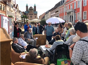Straßenaktion Zimmer auf der Straße 1a Auf dem Gehsteig der Maximilianstraße wurden Wohnzimmermöbel aufgestellt. Das Sofa und die Sessel sind besetzt mit Leuten, die zusammen diskutieren.