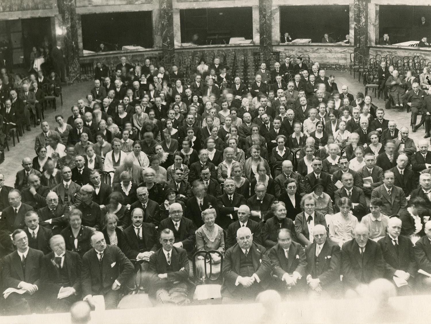 schwarz-weiß Fotografie vieler Menschen auf Stühlen sitzend in einem festlichen großen Saal.