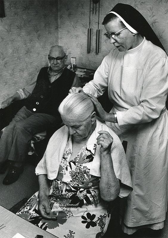 Ordensschwester steckt alter Dame die Haarfrisur. Im Hintergrund sitzt der Ehemann der Frau und schaut zu.