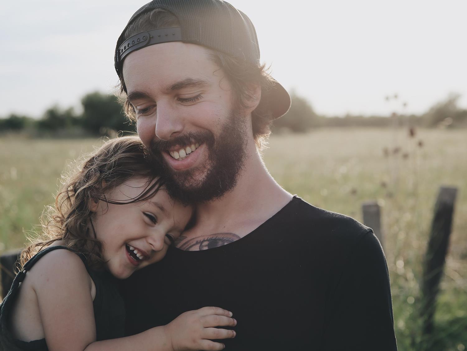 Ein junger Mann mit Vollbart umarmt ein kleines Mädchen - beide lächeln.