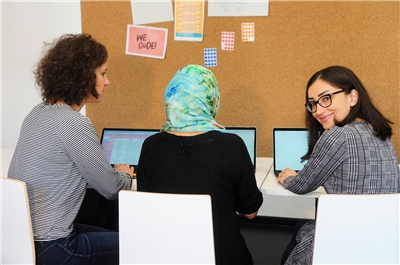Drei junge Frauen sitzen an einem Computer, eine von ihnen trägt ein Kopftuch.