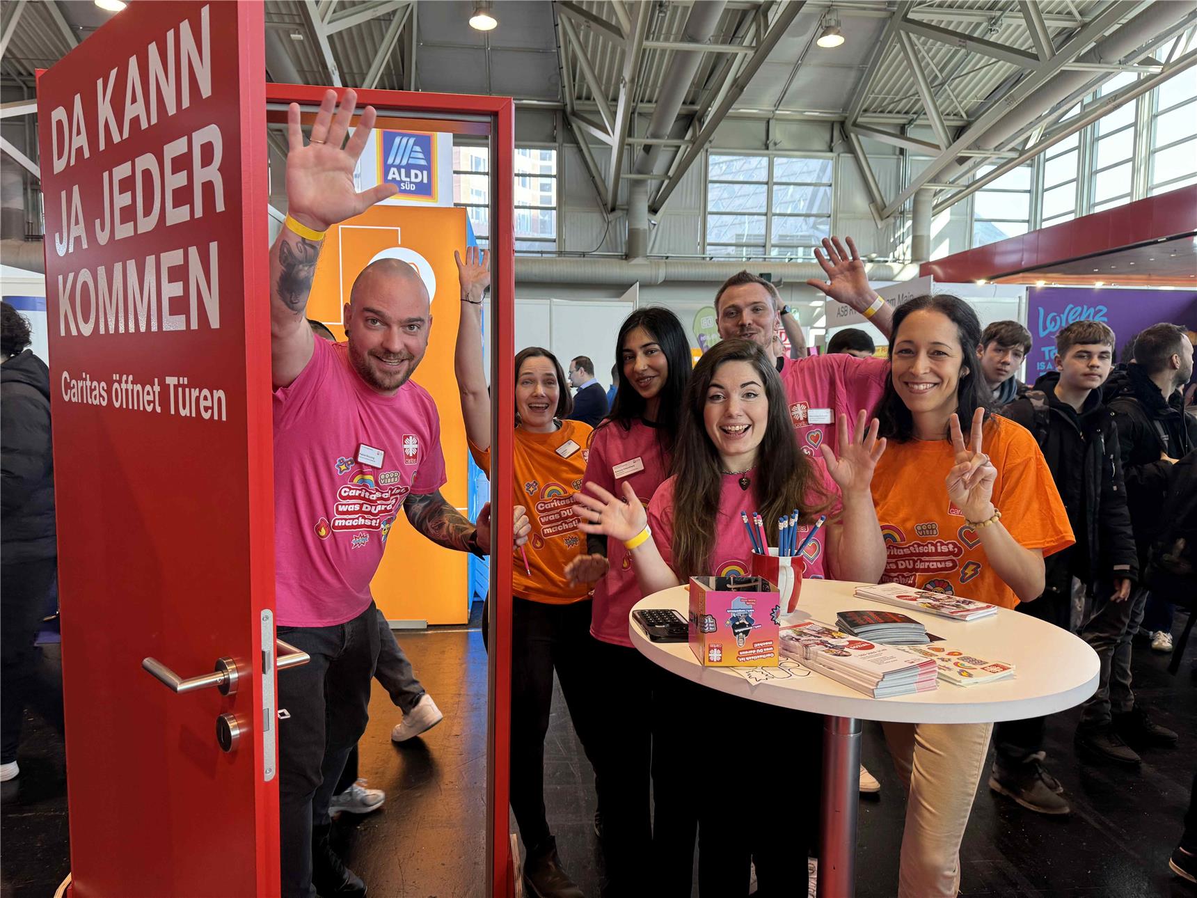 Die Caritas Mitarbeiter*innen akquirieren auf der Frankfurter Messe neue Mitarbeiter*innen und schauen lachend aus dem Türrahmen heraus.