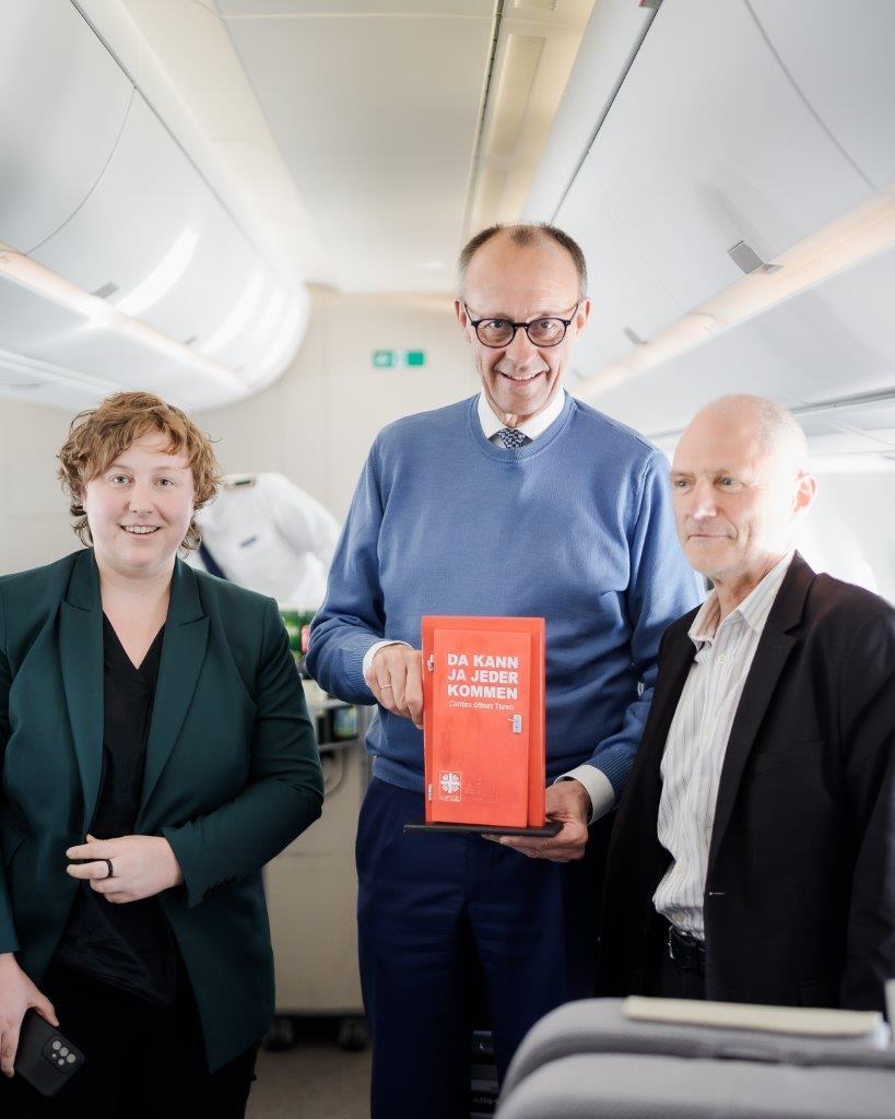 Paulina Hauser, Bundeskanzler Friedrich Merz, Christian Weickhardt in einem Flugzeug. Friedrich Merz hält ein kleines Modell einer roten Kampagnentür in Händen.