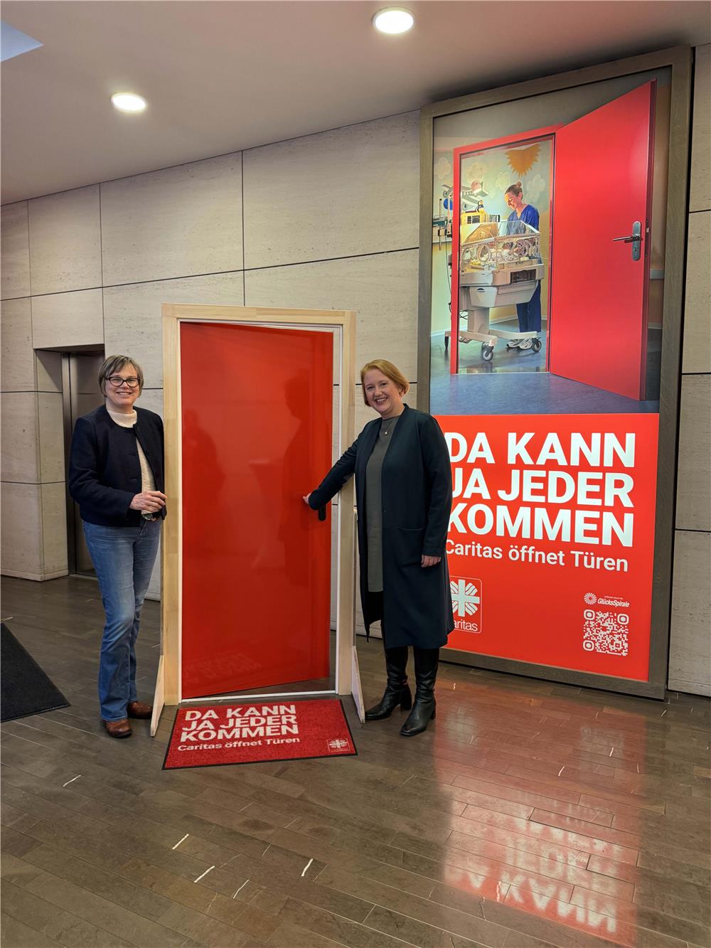 In einer schönen Aula stehen Caritas Präsidentin Frau Welskop-Deffa mit Lisa Paus, der Bundesministern für Familie, Senioren, Frauen und Jugend, vor einer roten Tür. Lisa Paus öffnet die Tür.