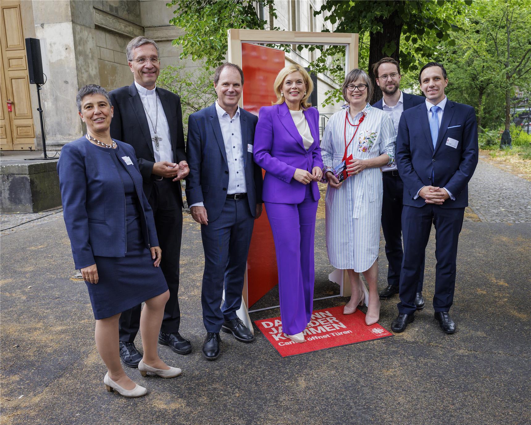 Die prominenten BesucherInnen stehen vor der roten Kampagnentür, die geöffnet ist. Die Szene spielt sich in einem Park ähnlichem Setting ab, neben der Kirche.