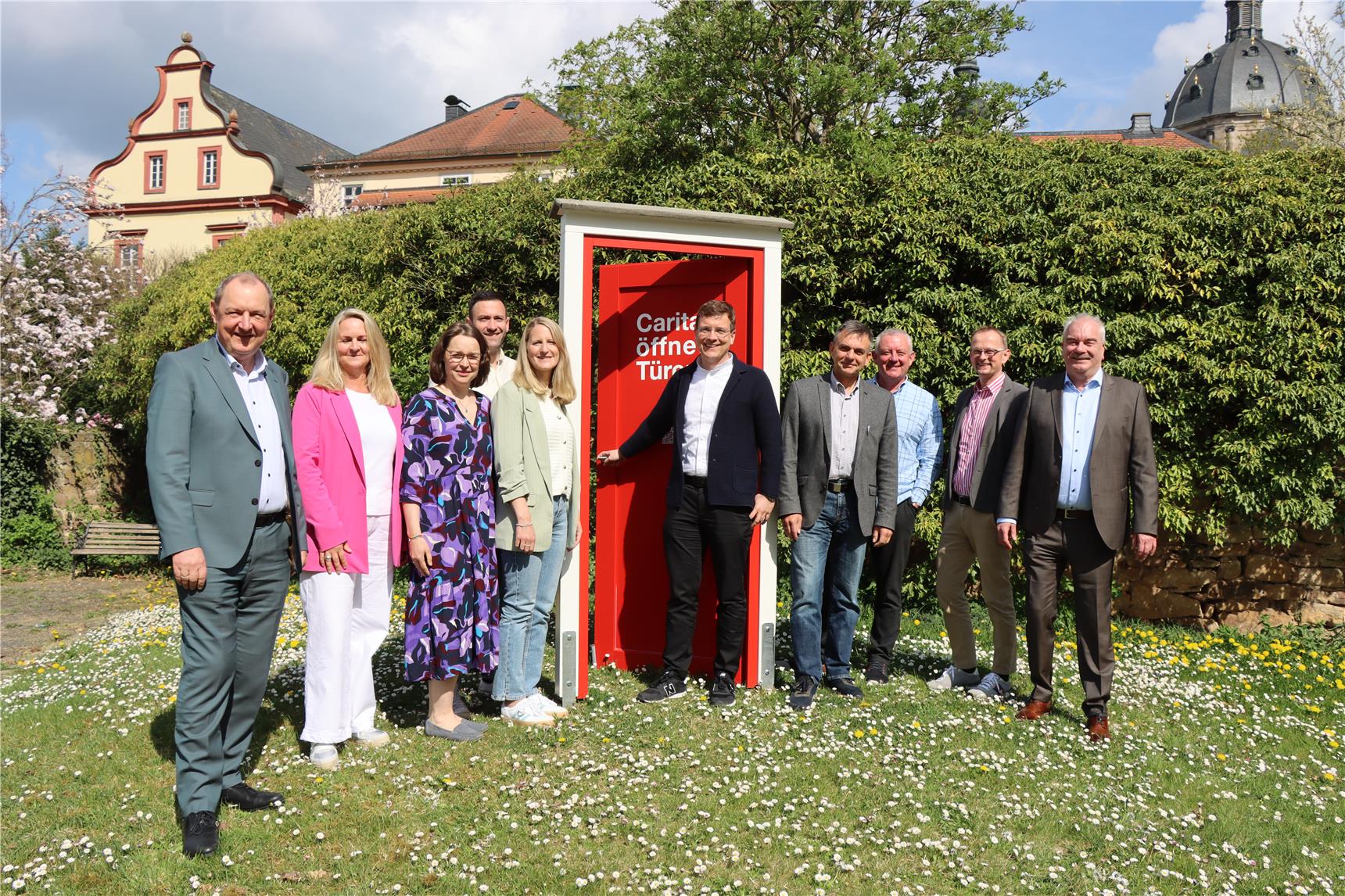 Der versammelte Caritasvorstand steht auf einer blühenden Frühlingswiese mit der roten Kampagnentür.
